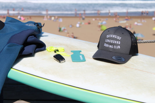 Tabla de surf en la playa con neopreno, llave electrónica de coche, KGUARD Key Pouch turquesa con cordón amarillo y gorra de surf, con el mar y surfistas al fondo.